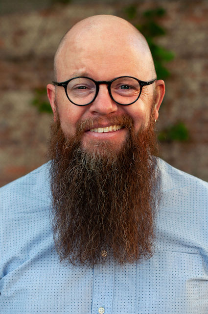 a portrait of a bald man with 
        a large red/brown beard and round glasses. 
        He is wearing overalls and a dark blue sweater, 
        and doing his best to smile in a professional sort of way.